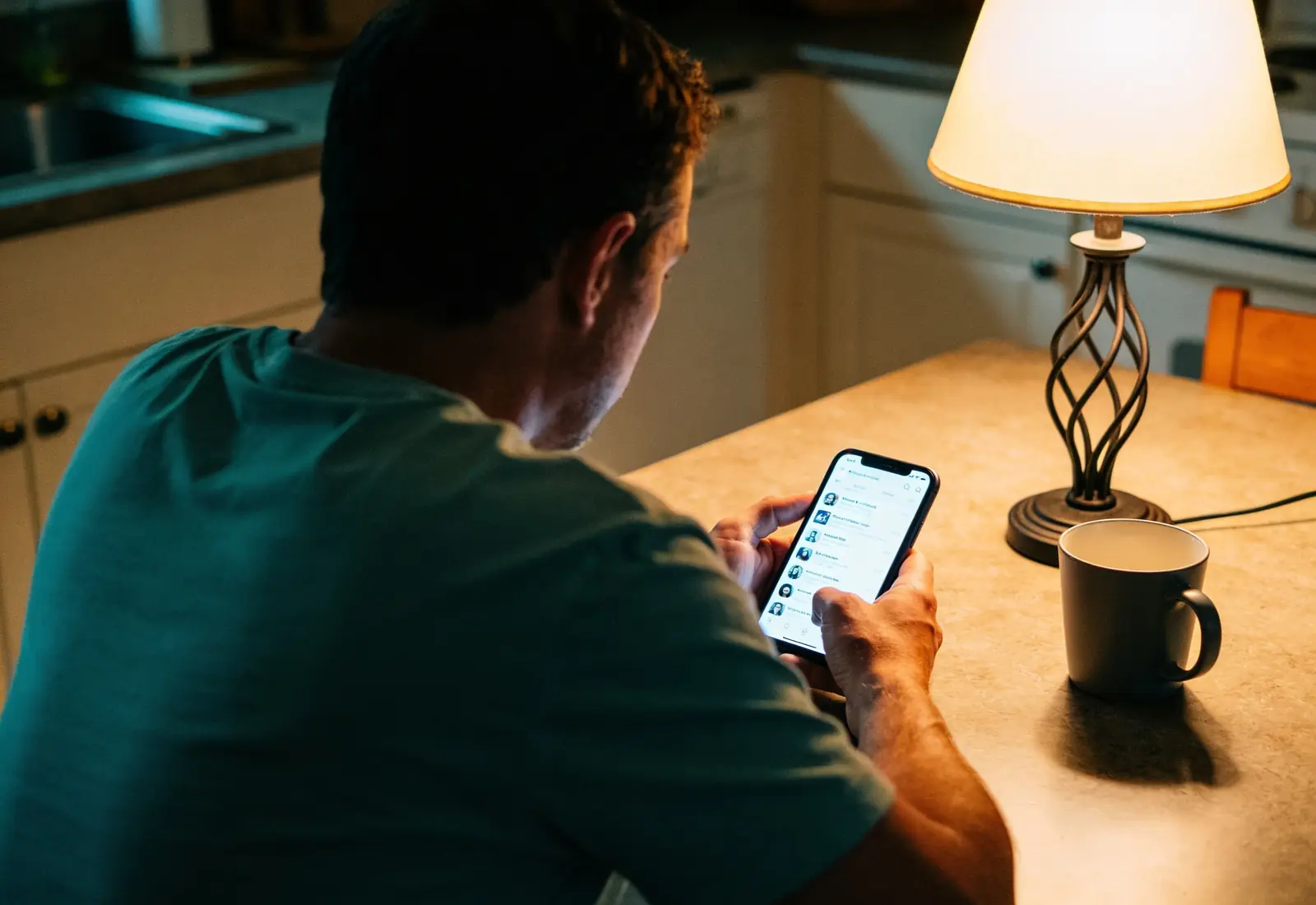 Homem de costas à mesa da cozinha à noite, lendo o celular com um feed de rede social desfocado, com um abajur e uma xícara ao lado