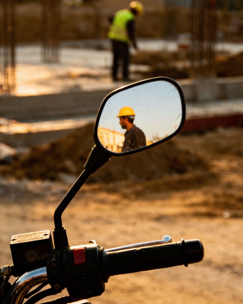 Retrovisor de moto refletindo um trabalhador de capacete num canteiro de obras