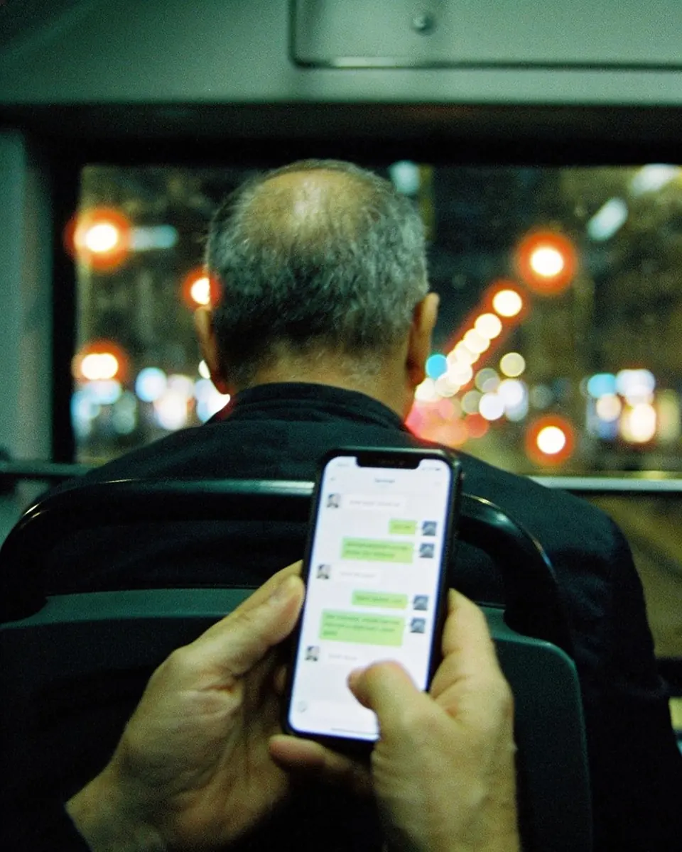 Homem de costas num ônibus à noite segurando o celular com as duas mãos, a tela acesa com uma conversa desfocada