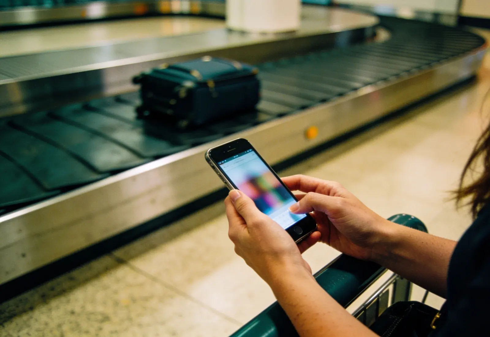 Mãos de uma mulher digitando no celular ao lado da esteira de bagagens, com uma única mala girando desfocada ao fundo