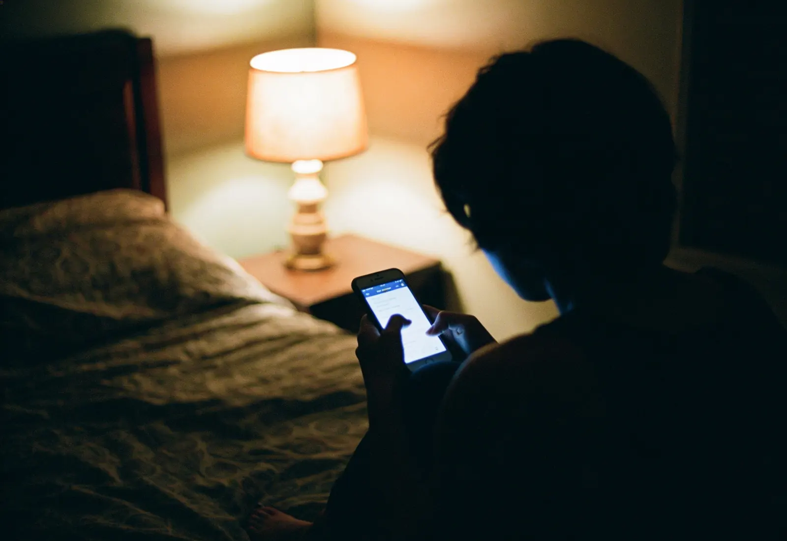 Pessoa de costas sentada na cama à noite, com a luz do celular iluminando as mãos