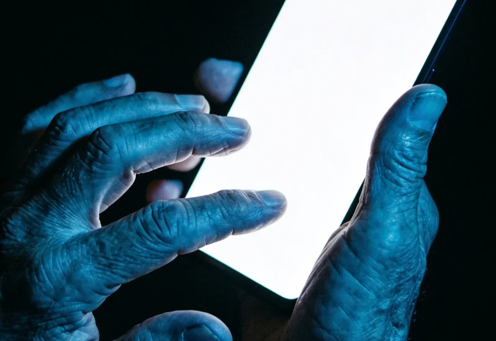Mãos de pessoa idosa digitando em celular no escuro, com a tela iluminando os dedos