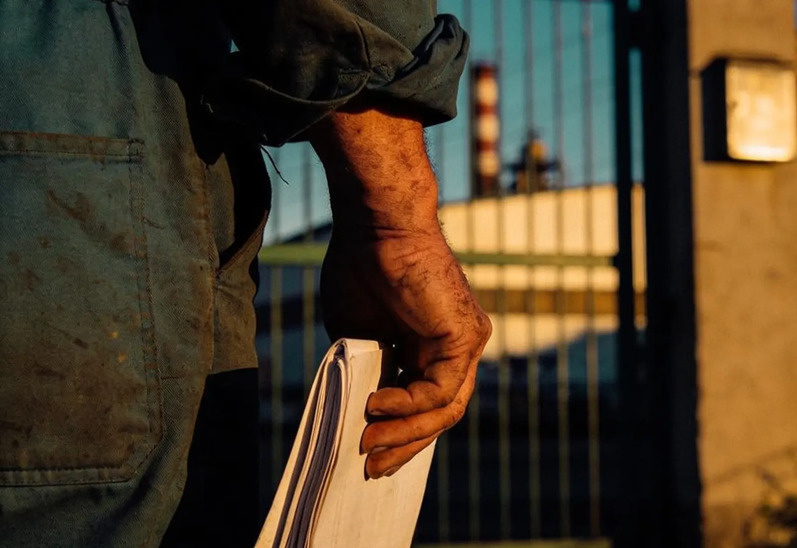 Mãos de trabalhador industrial segurando um documento dobrado diante do portão de uma fábrica ao entardecer