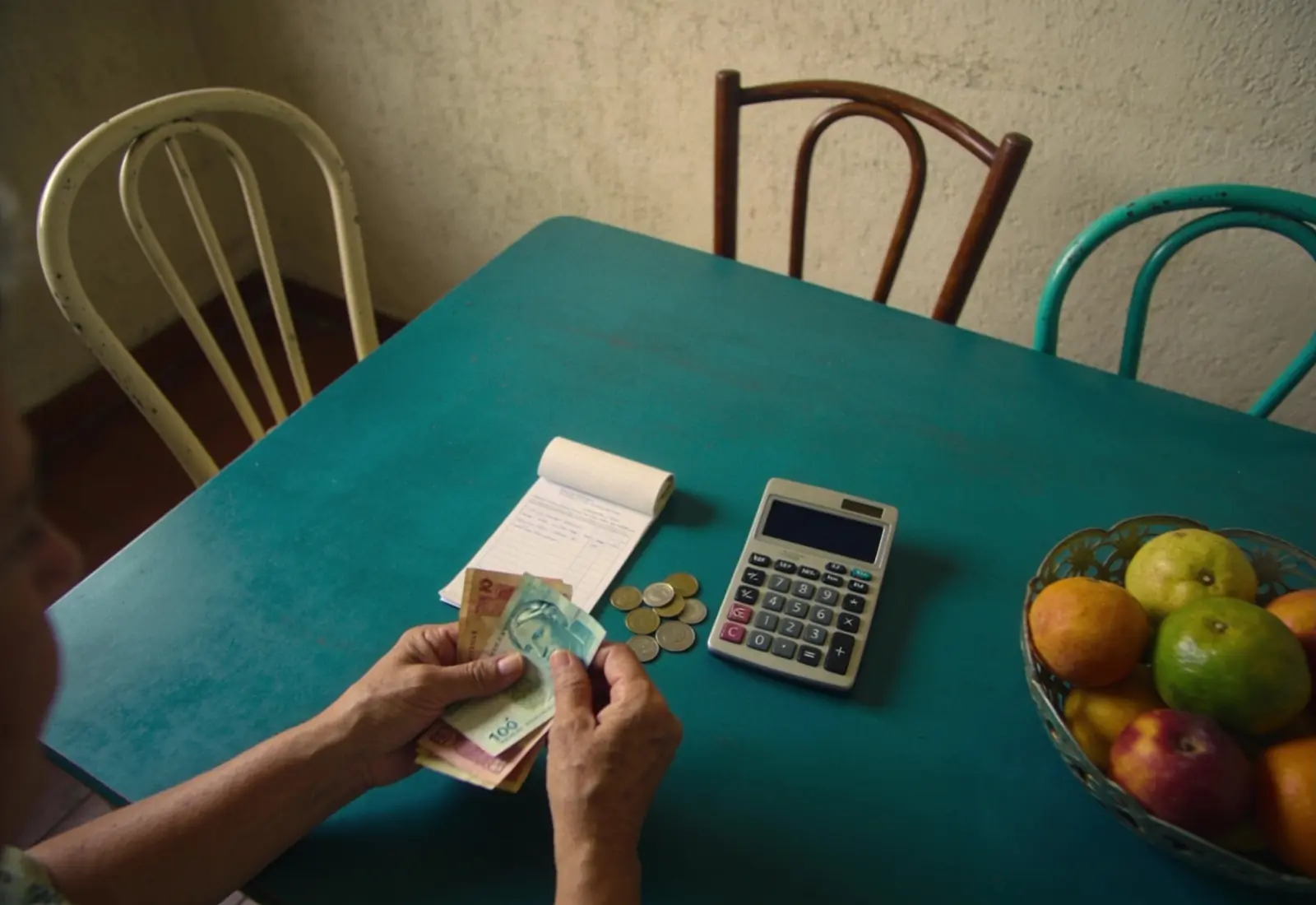 Mãos de uma mulher à mesa da cozinha separando cédulas e moedas ao lado de um documento dobrado e de uma calculadora