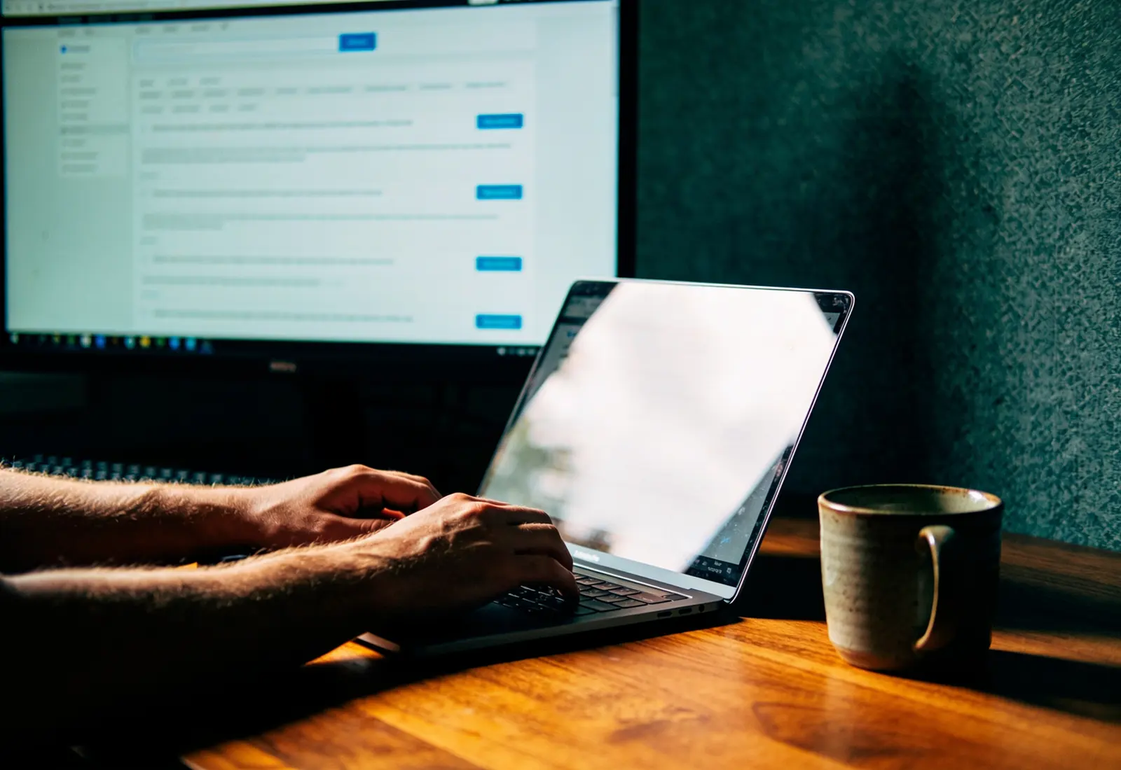 Mãos digitando num notebook ao lado de um monitor com uma página de busca desfocada