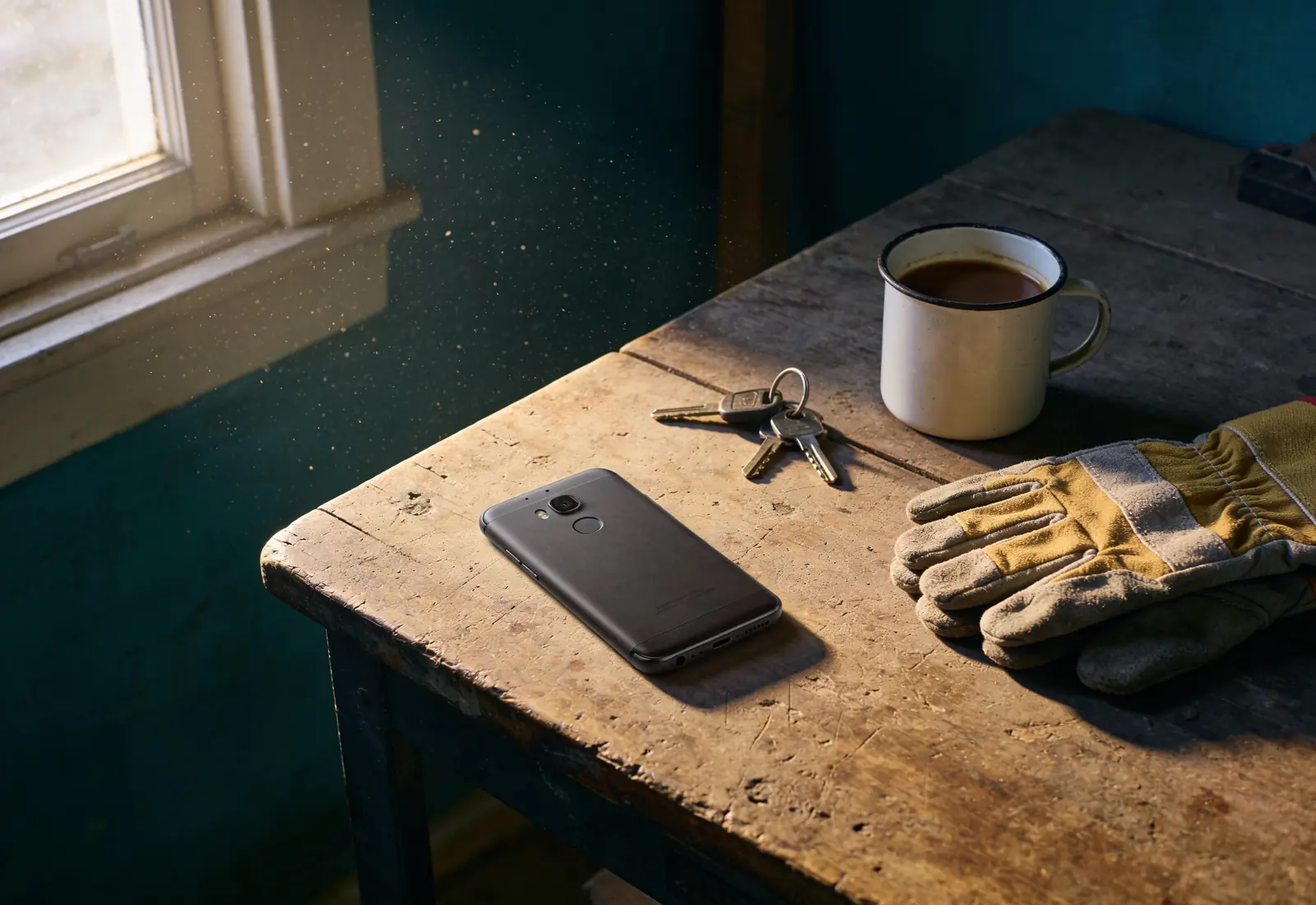 Celular virado para baixo numa mesa de trabalho de madeira, ao lado de um par de luvas, um molho de chaves e uma xícara de café pela metade